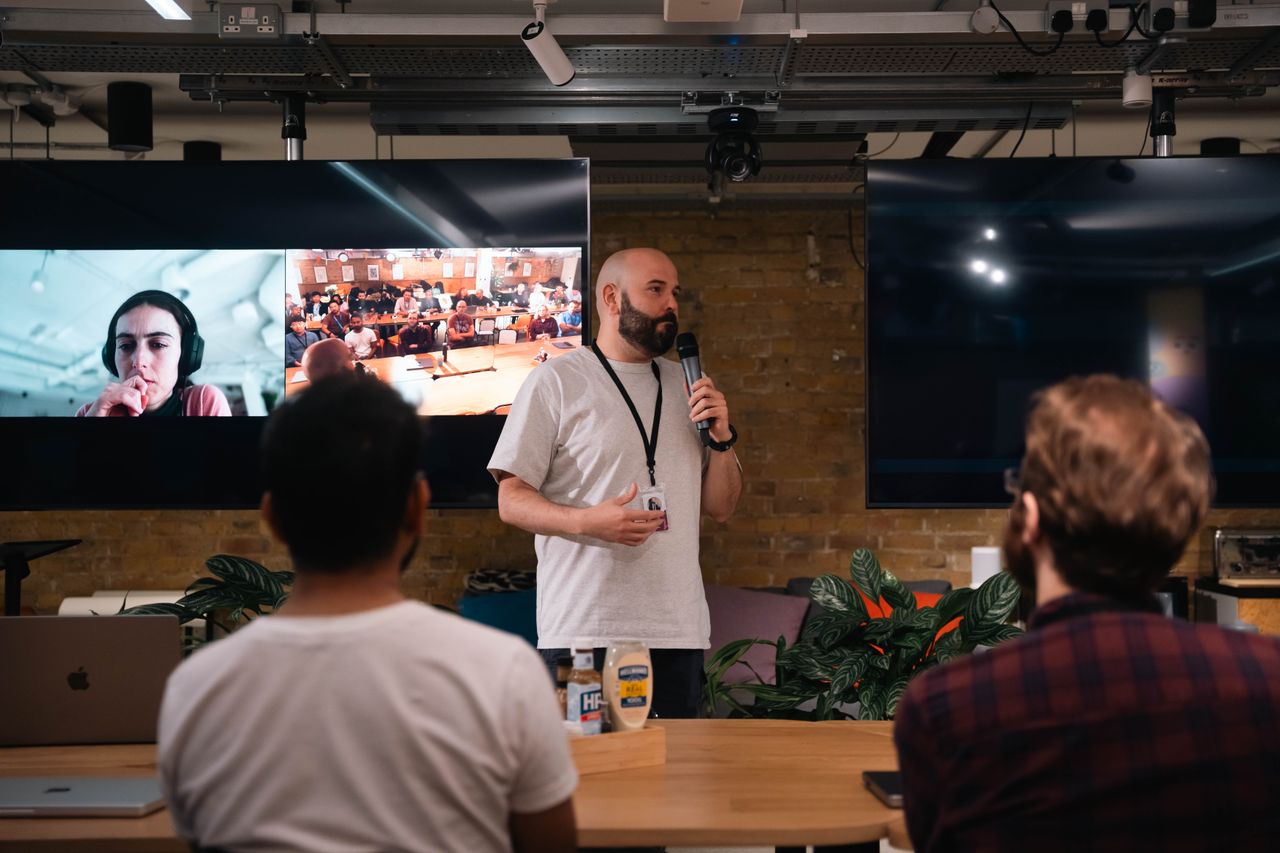 Speaker delivering a presentation in a contemporary office, with an audience and a virtual call displayed on screens.